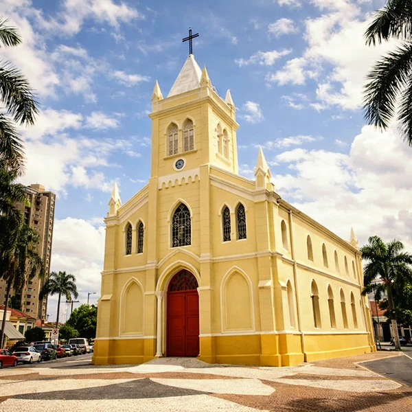 Igreja de estilo gótico com fachada amarela e torre com cruz ao topo, localizada em um bairro urbano com árvores e carros ao redor, sob céu parcialmente nublado.
