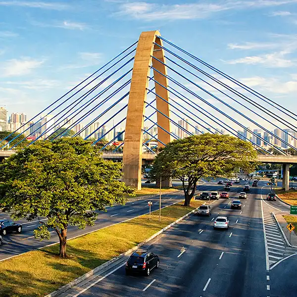 Imagem de uma ponte moderna sobre uma avenida movimentada na cidade, com árvores e prédios ao fundo, durante o dia ensolarado, destacando a arquitetura e o trânsito.