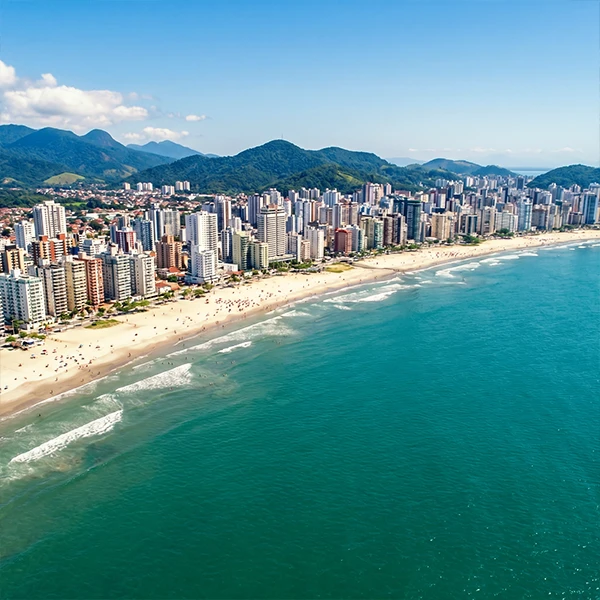 Vista aérea da orla de uma cidade costeira no Brasil, com muitos prédios altos próximo à praia, cercada por montanhas verdes ao fundo