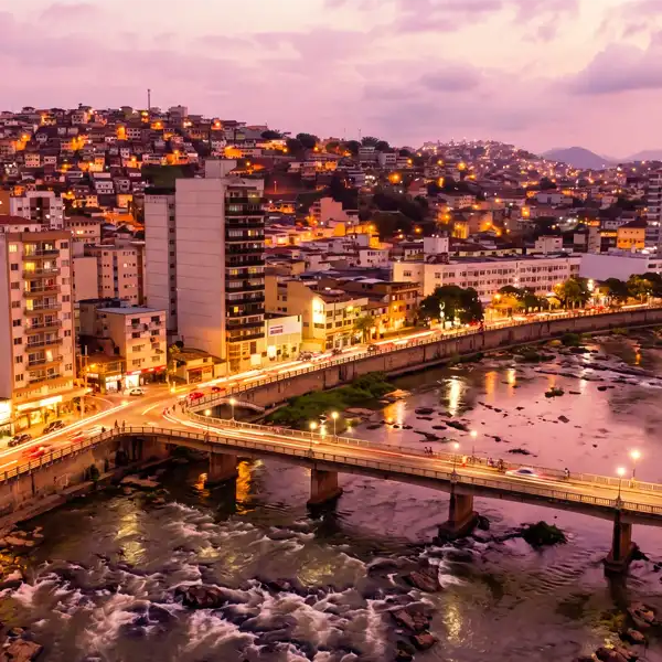 Imagem da cidade de Niterói ao entardecer, com lights brilhantes, ponte sobre o rio, bairros residenciais no morro e céu colorido ao pôr do sol, representando a beleza urbana e a vista panorâmica.