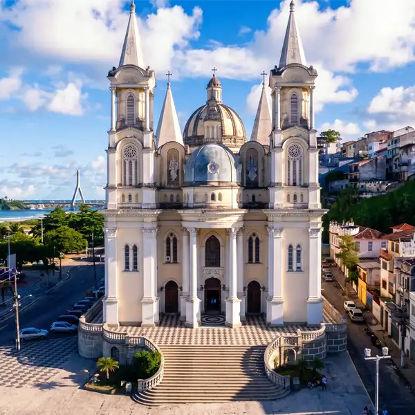 Imagem de uma imponente igreja com arquitetura clássica, torres altas e detalhes ornamentais, localizada em uma área urbana com vista para o rio e céu claro.