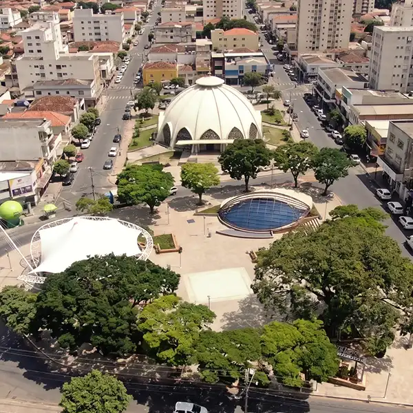 Imagem aérea de uma praça urbana com árvores, uma estrutura em forma de cúpula e um espaço com piscina, rodeada por ruas e edificações.