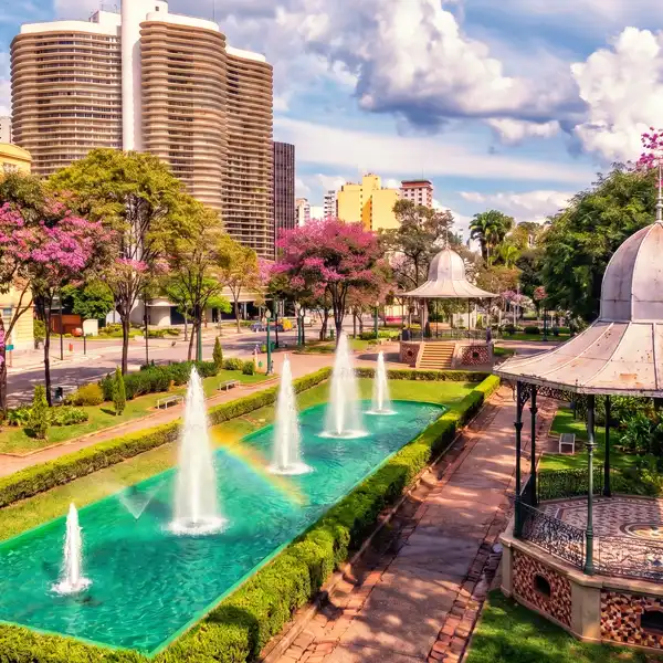 Parque urbano com fontes de água, árvores floridas e jardins bem cuidados em uma área central da cidade com prédios altos ao fundo e céu claro.