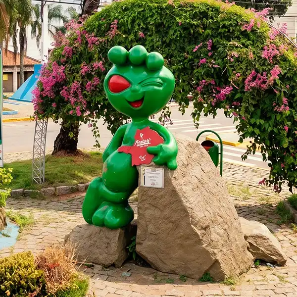 Estátua colorida de uma personagem infantil verde com detalhe vermelho, localizada em praça com árvores e flores ao fundo, celebrando cultura infantil.
