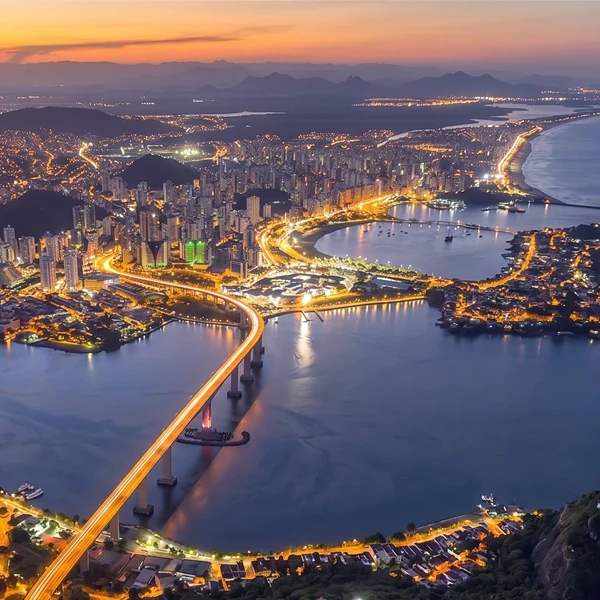 Vista aérea de uma cidade costeira durante o pôr do sol, com arranha-céus iluminados, pontes largas sobre o mar e montanhas ao fundo, mostrando a beleza urbana e natural.