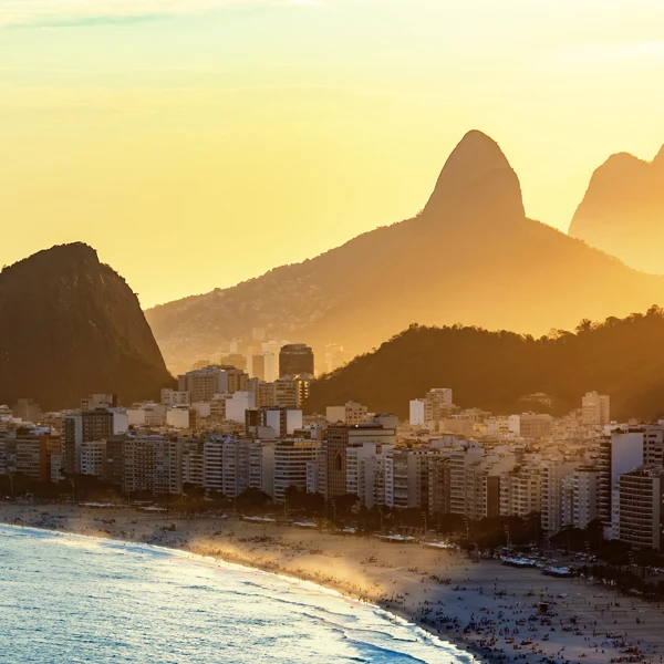 Vista panorâmica de uma cidade costeira no pôr do sol, com prédios altos à beira-mar e montanhas imponentes ao fundo, com praia e ondas ao primeiro plano.