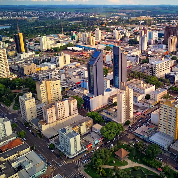 Vista aérea de uma cidade brasileira com prédios residenciais e comerciais, avenidas movimentadas e áreas verdes, ao entardecer.