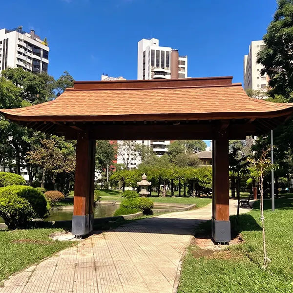 Gazebo de madeira com cobertura em formato curvo, assento central e bancos laterais, em um jardim com gramado, caminhos de piso e árvores, ao fundo prédios residenciais sob céu azul.