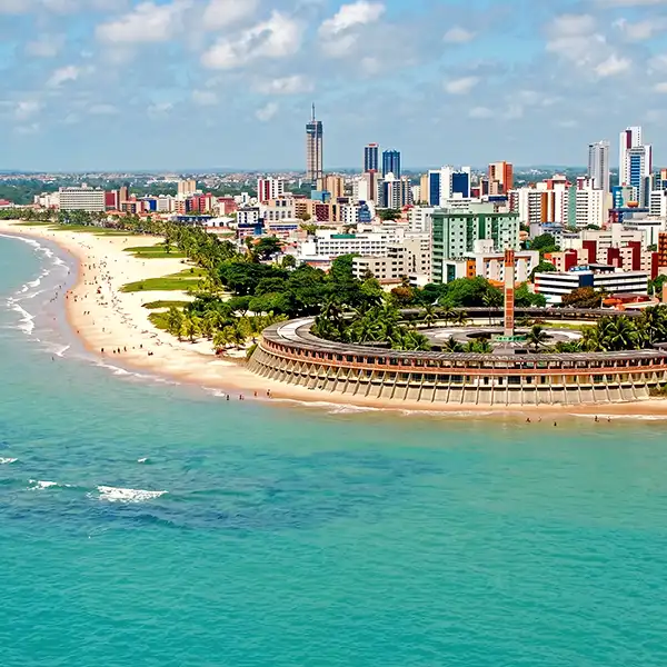 Vista panorâmica de uma cidade costeira com arranha-céus modernos, praia de areia branca e mar azul claro, em um dia ensolarado.