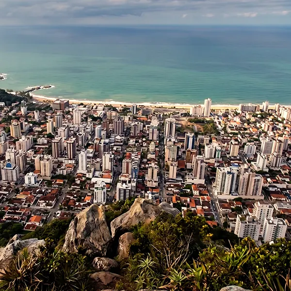 Vista panorâmica de uma cidade costeira com muitos prédios, casas e avenidas, além de pedras e vegetação em primeiro plano e o mar aberto ao fundo sob céu parcialmente nublado.