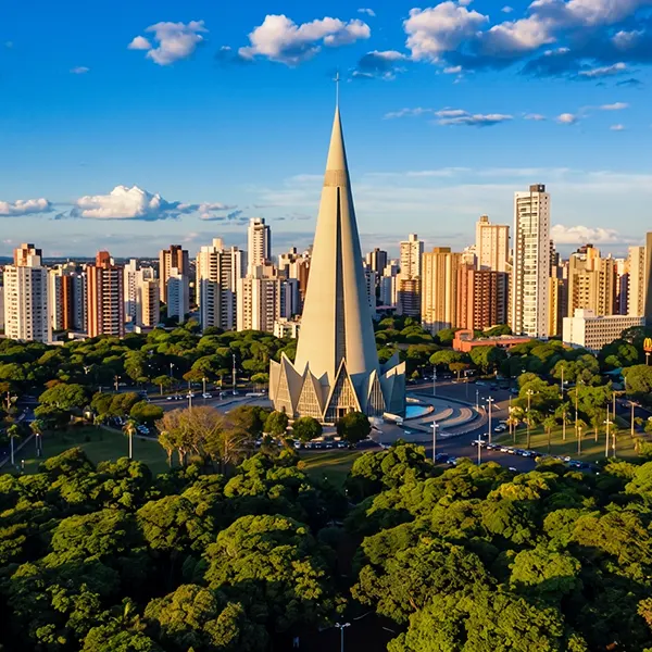 Vista aérea de uma cidade com arranha-céus ao fundo e um monumento arquitetônico em forma de torre pontiaguda no centro, cercado por árvores e vias ao entardecer, céu azul com nuvens.