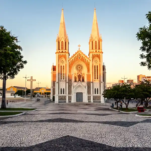 Catedral de pedra com duas torres altas sob um céu azul ao entardecer, localizada em uma praça com calçada de pedra, árvores ao redor e algumas pessoas caminhando