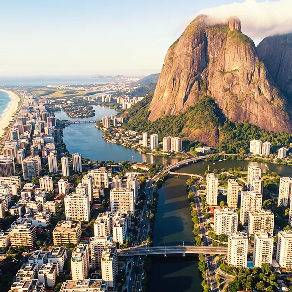 Vista aérea de uma cidade costeira com mar e um grande morro ao fundo, ao lado de um rio largo que atravessa o centro urbano e forma uma baía, com prédios residenciais e ponte