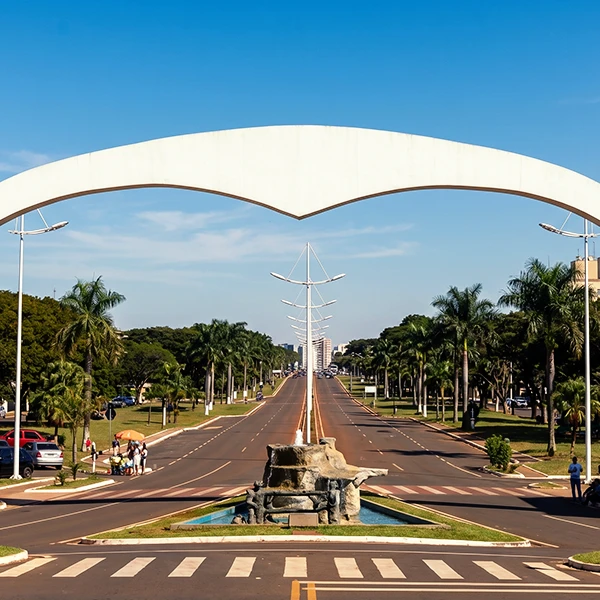 Vista de uma avenida larga e arborizada ao lado de palmeiras, com faixa de pedestres e canteiro central. Ao fundo, um arco branco monumental sobre a pista e um céu azul claro.