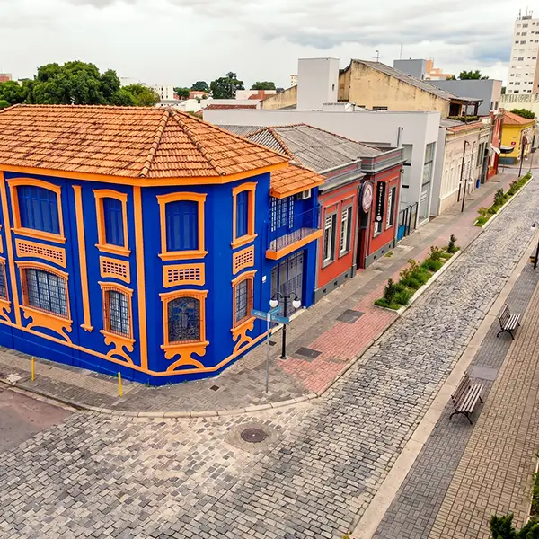 Vista aérea de rua com casas históricas coloridas, fachada azul e laranja com janelas e telhado de cerâmica, calçada de pedras e vegetação ao longo do caminho.