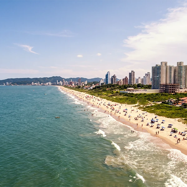 Vista aérea de uma praia urbana com areia clara e grande extensão de mar, ondas chegando à faixa de areia e pessoas tomando sol; ao fundo, prédios e arranha-céus sob céu azul com nuvens