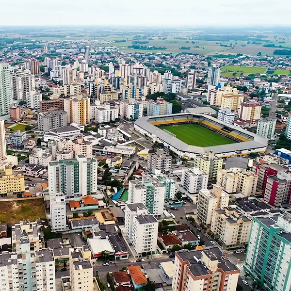Vista aérea de um bairro residencial com prédios e apartamentos, ruas internas e campo de futebol em primeiro plano, com áreas verdes e chácaras ao redor.