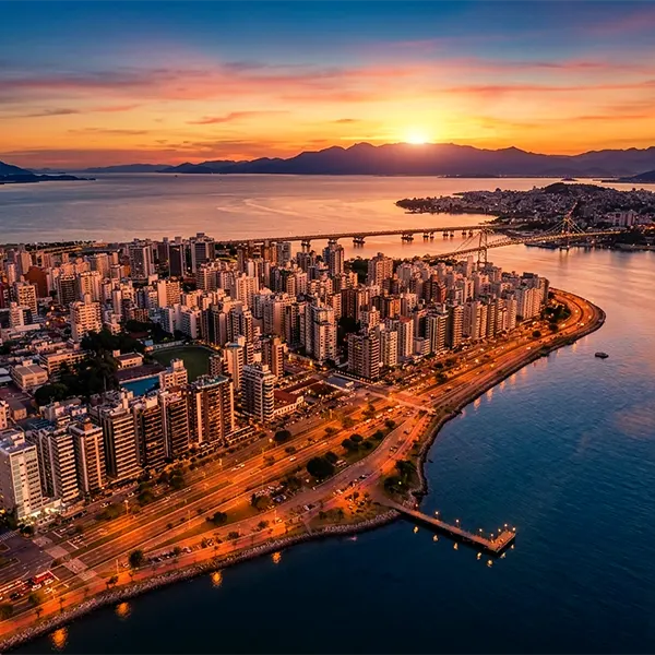 Vista aérea ao pôr do sol de uma orla urbana com prédios residenciais, ponte e cais ao longo da baía, com montanhas ao fundo e água ao centro da paisagem.