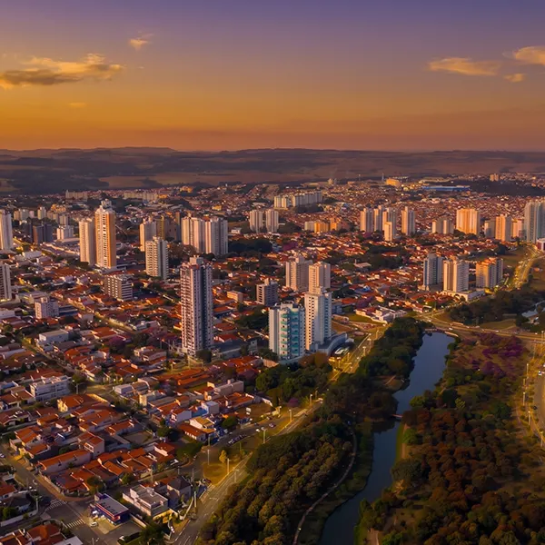 Vista aérea de uma cidade brasileira ao entardecer, com prédios residenciais altos, ruas iluminadas, casas e um rio cercado por áreas verdes, céu em tons de laranja e roxo.
