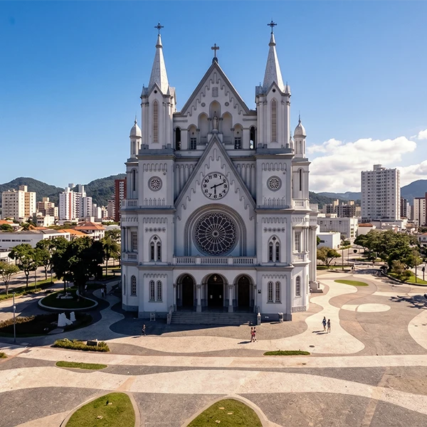 Vista aérea de uma igreja neogótica branca com torres e cruzes, grande fachada central com janelões em arco e relógio, cercada por praça ajardinada com calçadas e bancos, sob céu azul