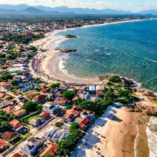 Vista aérea de uma praia com mar azul, faixa de areia e casas ao longo da orla, com pessoas na praia e vegetação no litoral ao fundo.
