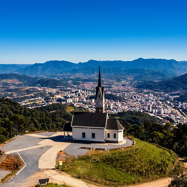 Capela no topo de uma colina com arquitetura branca e torre central, vista aérea ao pôr do sol, com uma cidade ao fundo e montanhas ao horizonte sob céu azul.